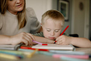 Boy with down syndrome drawing in book by mother at home - MASF40327