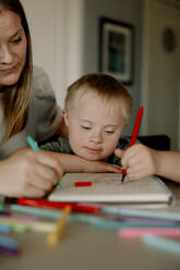 Boy with down syndrome drawing in book by mother at dining table - MASF40328