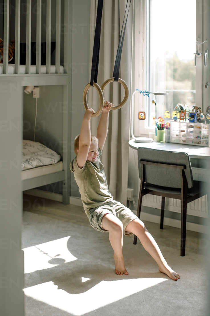 full length of happy boy hanging from gymnastic rings in bedroom at home MASF40329