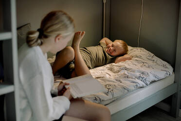 Sister reading picture book while sitting near brother with down syndrome lying on bed at home - MASF40330