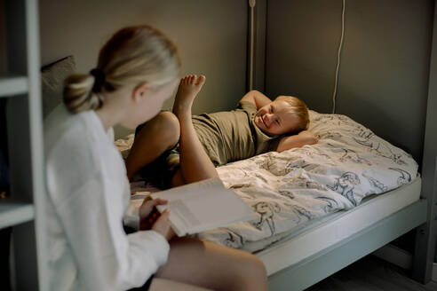 Sister reading picture book while sitting near brother with down syndrome lying on bed at home - MASF40330