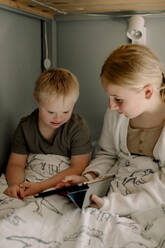 Girl sharing digital tablet with brother sitting on bed at home - MASF40332