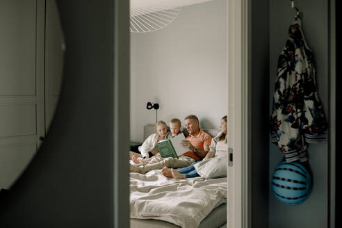 Man reading picture book for children while sitting on bed seen through doorway at home - MASF40335