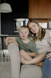 Portrait of mother and son with down syndrome sitting together on sofa at home - MASF40339