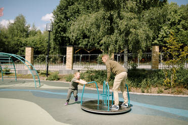 Boy with down syndrome spinning father on carousel at park during sunny day - MASF40348