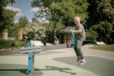 Full length side view of happy boy with down syndrome enjoying seesaw at park - MASF40351