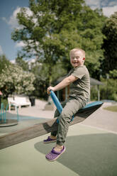 Volle Länge Seitenansicht Porträt des glücklichen Jungen mit Down-Syndrom sitzt auf Wippe im Park - MASF40353