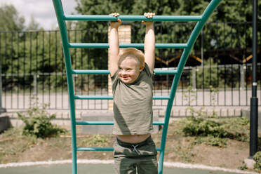 Smiling boy with down syndrome hanging from jungle gym at park - MASF40357