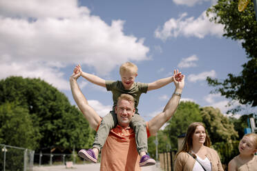 Father carrying son on shoulders while walking by woman and daughter at park during sunny day - MASF40359