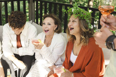 Cheerful women with drinks sitting by young man at patio in back yard - MASF40375