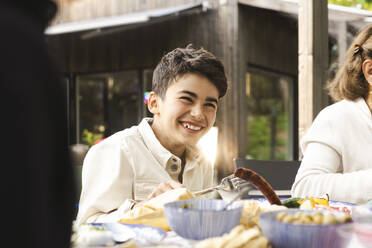 Cheerful boy sitting at dining table in patio during dinner party - MASF40391