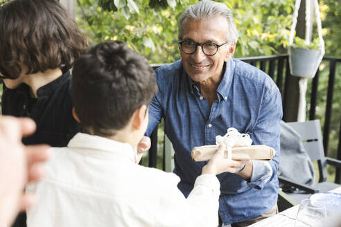 Smiling senior man giving gift to grandson during dinner party - MASF40393