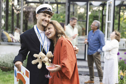 Smiling mother and son embracing while standing at back yard during summer party - MASF40415