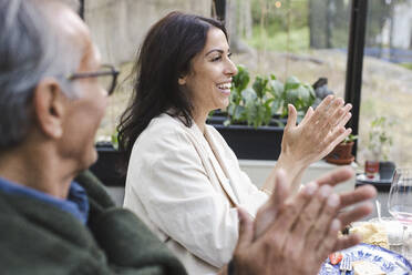 Side view of happy man and woman clapping while sitting at patio during summer party - MASF40435