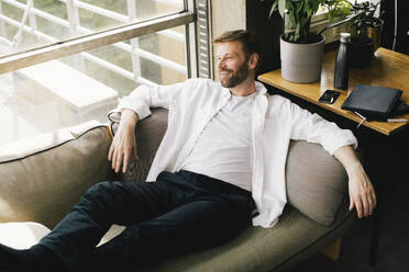 High angle view of smiling thoughtful mature businessman reclining on sofa at office - MASF40447