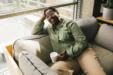 Smiling businesswoman sitting with coffee cup while day dreaming at office - MASF40451