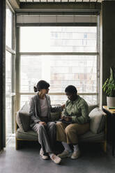 Full length of mature businesswoman consoling female colleague sitting on sofa against window at office - MASF40454