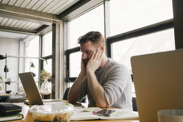 Confused businessman sitting with head in hands looking at laptop on desk - MASF40480