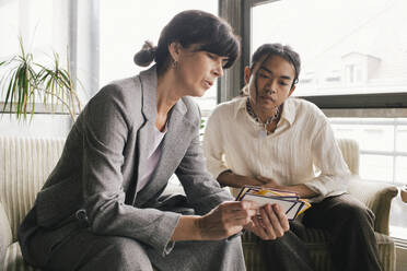 Businesswoman discussing over placard with non-binary person while sitting at office - MASF40486