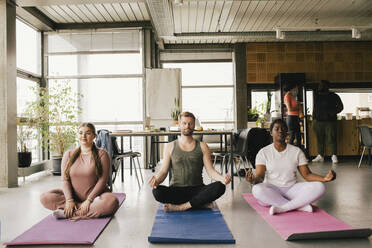 Full length of multiracial professionals meditating together on mats at office - MASF40491