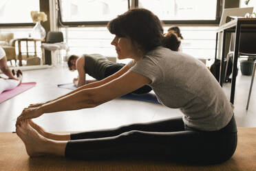 Full length side view of flexible mature businesswoman touching toes while practicing yoga on mat at workplace - MASF40503