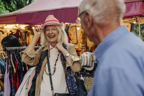 Cheerful senior woman trying on hat by man standing at market - MASF40525