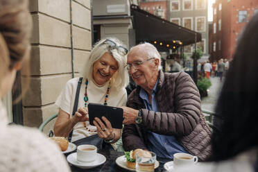 Happy senior couple sharing smart phone while sitting at sidewalk cafe - MASF40538