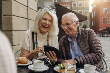 Cheerful senior couple sharing smart phone while sitting at sidewalk cafe - MASF40539