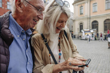 Happy senior couple sharing smart phone while exploring city - MASF40546