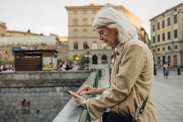 Side view of senior woman leaning on railing while using smart phone in city - MASF40561