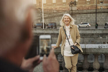 Senior man photographing happy woman leaning on railing - MASF40562