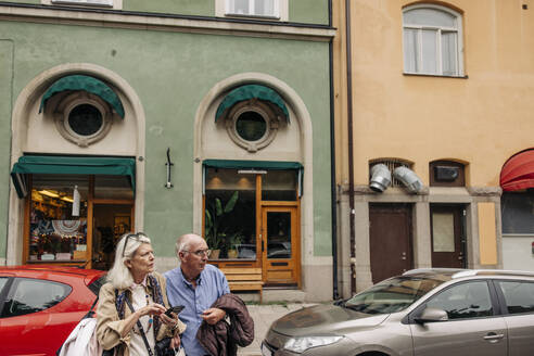 Confused senior man and woman looking away while crossing against building - MASF40578