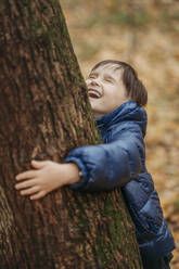 Glücklicher Junge, der einen Baum im Herbstpark umarmt - ANAF02384