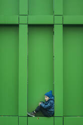 Boy in warm clothing sitting near green wall - ANAF02386