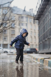 Niedlicher Junge spielt auf nasser Straße nach Regen - ANAF02388