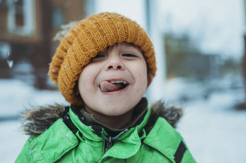 Playful boy wearing warm clothes sticking out tongue in winter - ANAF02395