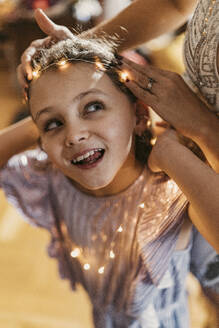Woman adjusting string light on daughter's forehead at home - MFF09454