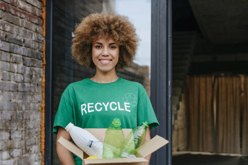 Smiling activist holding cardboard box with plastic bottles - YTF01296