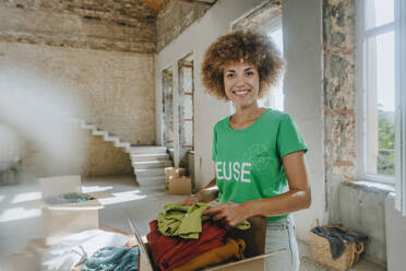 Lächelnde Aktivistin mit lockigem Haar stapelt Kleidung in einer Pappschachtel in ihrer Wohnung - YTF01302