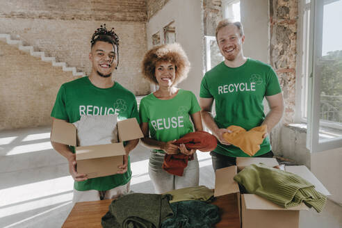 Smiling activists holding secondhand clothes with cardboard boxes in apartment - YTF01316