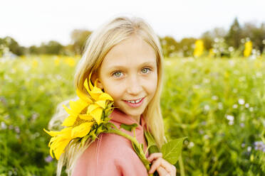 Lächelndes blondes Mädchen hält Sonnenblume im Feld - NJAF00604