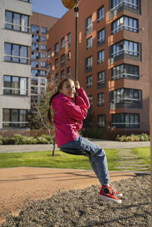 Happy girl playing on swing in front of buildings at playground - LESF00503