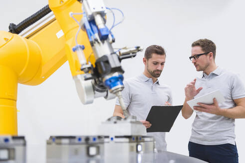 Ingenieure mit Tablet-PC und Aktenordner bei einer Diskussion in der Nähe eines Roboterarms in einer Fabrik - DIGF20953