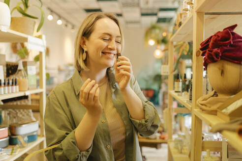 Smiling woman with eyes closed smelling face oil in zero waste store - VIVF01177