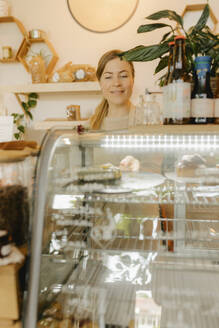 Lächelnde Kellnerin arrangiert frischen Kuchen im Kühlschrank auf dem Tresen eines Cafés - VIVF01182