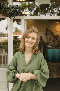 Smiling blond woman with hands clasped standing outside cafe - VIVF01237