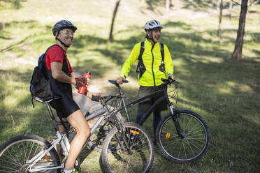 Lächelnder Mann, der eine Wasserflasche hält und neben einem Freund mit Fahrrad im Wald steht - JCCMF10885