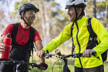 Glücklicher Mann steht mit Freund in der Nähe von Fahrrad und hält Wasserflasche im Wald - JCCMF10886