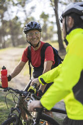 Smiling man holding water bottle and talking to friend in forest - JCCMF10887