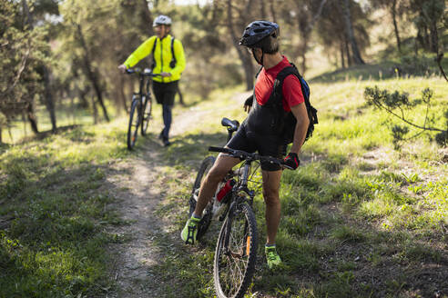 Mature man standing with bicycle waiting for friend at forest - JCCMF10889